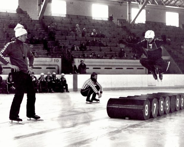 Photographie en noir et blanc où l'on voit trois hommes sur une patinoire. L'un d'eux est en train de sauter par-dessus des barils.