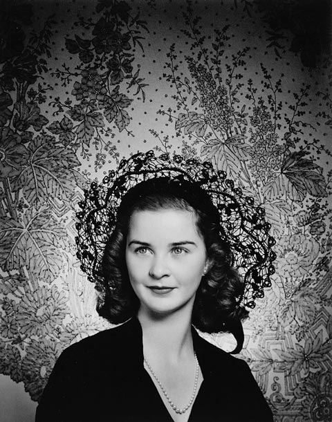 Photographie en noir et blanc d'une femme portant un haut noir, un chapeau et un collier. Elle se trouve devant une tapisserie florale.