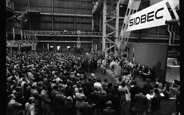 La photo a été prise à l’intérieur de l’usine Sidbec lors de son inauguration. Sur une scène aménagée pour l’occasion, on voit plusieurs dignitaires, dont le premier ministre Robert Bourassa au micro. Au parterre, des centaines de personnes sont rassemblées pour assister à l’événement.