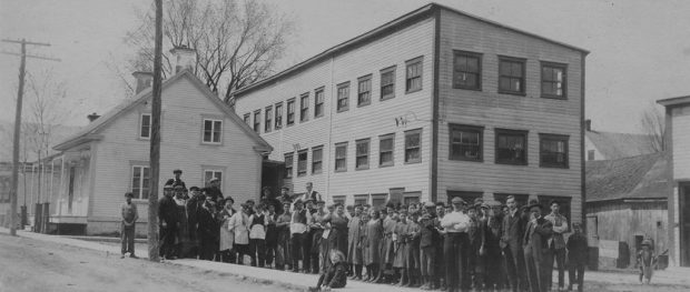 La photo montre les employés rassemblés devant la manufacture Charron. Il s’agit d’une bâtisse en bois de trois étages, construite à l’arrière d’une maison sur la rue St-Antoine.