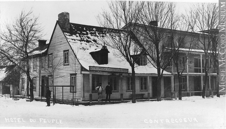 Voici une photographie de l’Hôtel du Peuple, prise vers 1910. On y voit qu’un deuxième bâtiment construit sur trois étages a été annexé à la maison en bois blanche sur laquelle l’enseigne a été apposée. Sur l’enseigne, on peut lire : Hôtel du Peuple F de S Gervais — Prop.