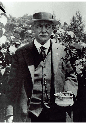 Photo de Joseph Papin II habillé en complet-veston dans une cour extérieure. Il porte un chapeau et semble tenir un bol de fruits avec sa main gauche.