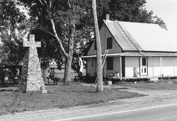 Cette photo montre une petite maison en bois blanche située au bord du fleuve Saint-Laurent. Juste à côté est érigée une croix en pierre désignant le lieu où aurait été construite la première église de Contrecœur.  