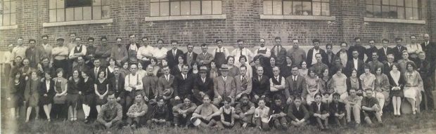La photo, prise devant l’usine, montre les employés de la manufacture d’Albert Charron en 1928.  Ils sont près d'une centaine; hommes, femmes et enfants.  On voit le mur de brique et les grandes fenêtres à l'arrière.