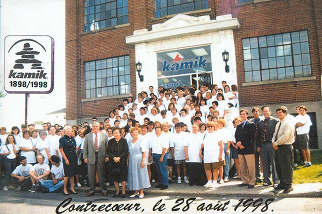 Sur cette photo, on voit tous les employés de la manufacture Kamik réunis devant l’usine à l’occasion du 100e anniversaire de l’entreprise.  Ils sont plus d’une centaine, principalement des femmes, et portent tous un chandail blanc.  À l’avant et sur le côté droit, on voit des membres de l’équipe de direction.