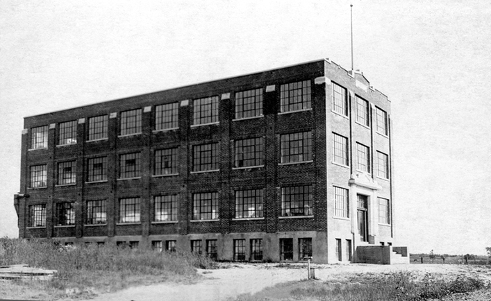 La photo montre la Manufacture Charron, peu de temps après sa construction en 1924. Un chemin de terre mène à l’entrée principale.  On ne voit aucun autre bâtiment autour.  C’est une construction en brique de quatre étages qui semble située au milieu d’un champ.  