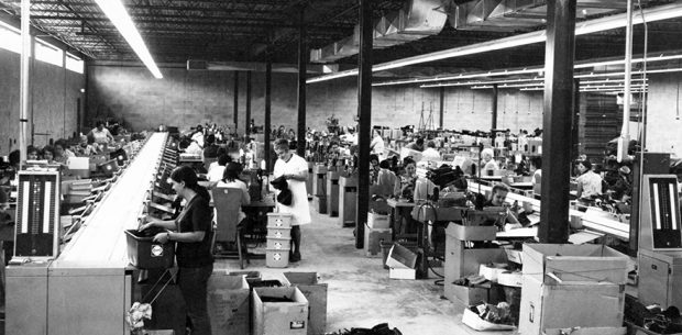 La photo montre le département de couture. On voit une quarantaine d’ouvrières installées de chaque côté de deux longs convoyeurs qui traversent la pièce.