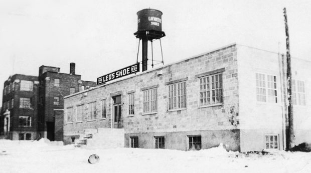 Photo montrant la manufacture Leo’s shoe Reg’d. C'est une bâtisse en brique grise de deux étages. On y voit aussi à l’arrière la manufacture Lafayette et son château d’eau.