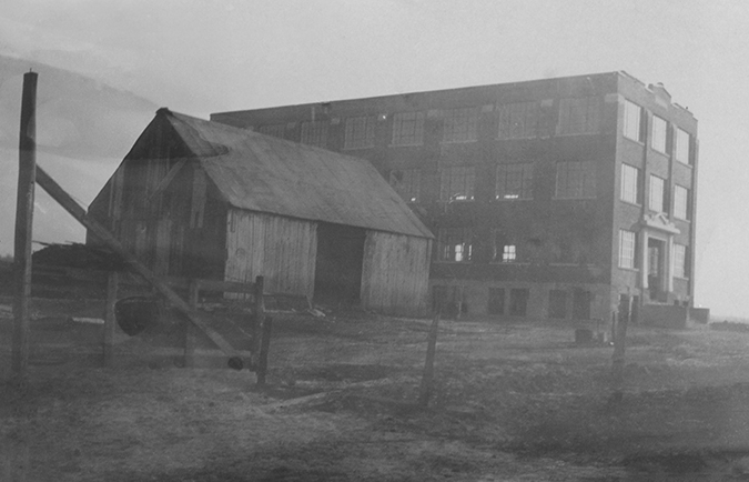 La photo montre la Manufacture Charron, peu de temps après sa construction en 1924. Il s’agit d’une grande bâtisse en briques de quatre étages. Il n’y a même pas encore de rue qui passe devant. On voit seulement une grange en bois à côté de l’usine. 