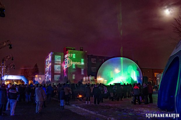 Cette photo montre l’usine Lafayette éclairée par des projecteurs lors des célébrations du 350e anniversaire de la ville de Contrecœur. On voit aussi un dôme futuriste devant l’ancienne manufacture et des centaines de personnes rassemblées.