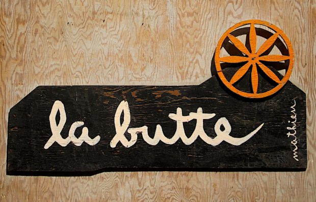 Photo couleur de l’enseigne de La Butte en contreplaqué peint surmonté d’une petite roue de charrette orange.