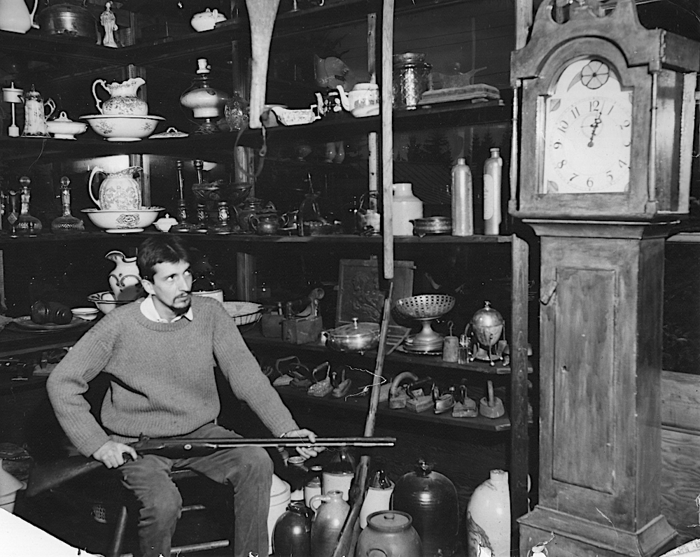 Photo noir et blanc du magasin d’antiquité de la Butte. Gilles Mathieu est assis et tient un vieux fusil. Une horloge grand-père prédomine devant des tablettes sur lesquels sont présentées des antiquités.