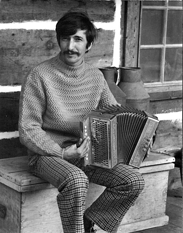 Photo noir et blanc de Gilles Mathieu assis sur un banc de bois sur le balcon de sa maison pièce sur pièce sur le site de La Butte et jouant de l’accordéon.