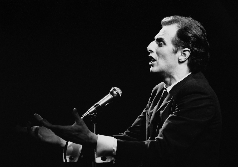 Photo noir et blanc de Gilles Vigneault bouche ouverte et mains tendues devant son micro.