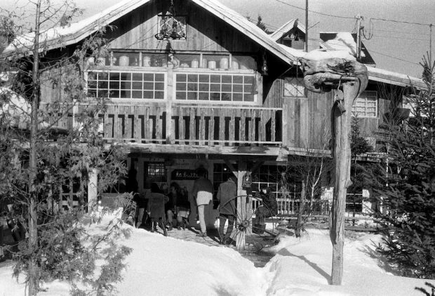 Photo noir et blanc de l’entrée principale de La Butte l’hiver à l’extérieur. Sur ses murs de bois de grange, une grande galerie longe des fenêtres à carreaux.