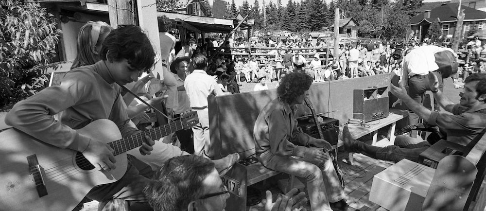 Photo noir et blanc d’une foule extérieure compacte aux Fêtes de la Saint-Jean organisées à La Butte. À l’avant-plan, un jeune homme joue de la guitare.