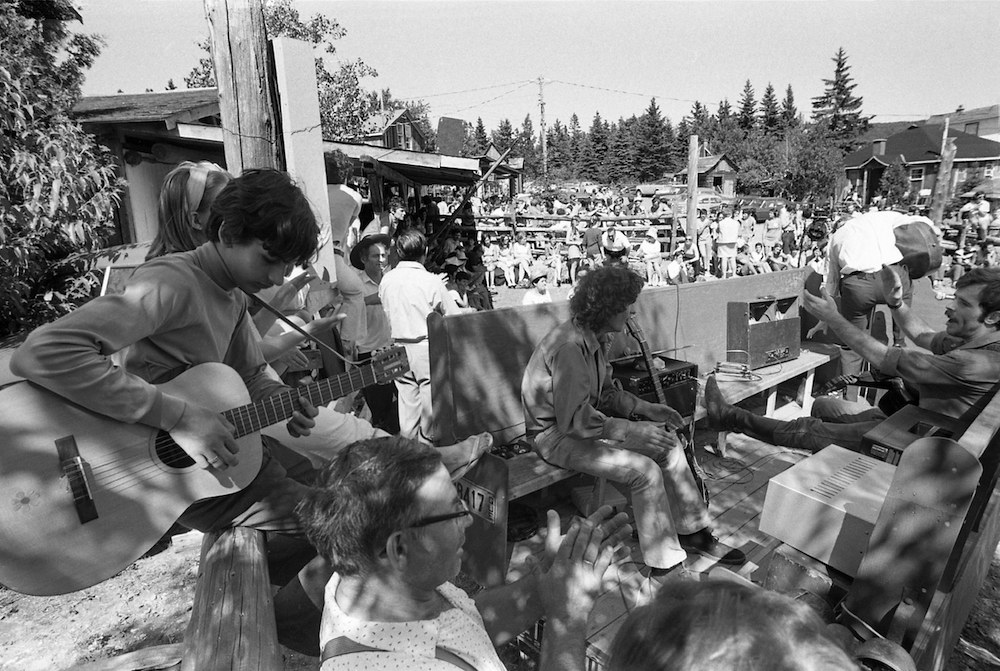 Photo noir et blanc d’une foule extérieure compacte aux Fêtes de la Saint-Jean organisées à La Butte. À l’avant-plan, un jeune homme joue de la guitare.