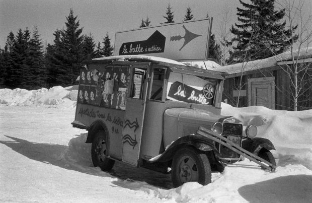 Photo noir et blanc d’un vieux camion de livraison des années 1930 sur lequel sont installées des affiches indiquant la direction à prendre pour se rendre à La Butte. 