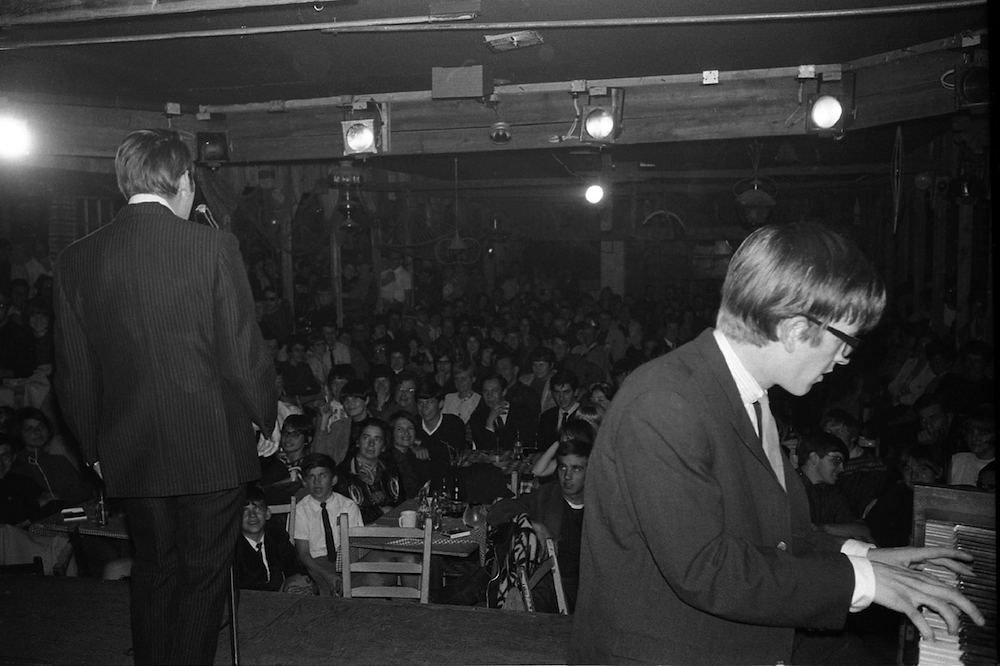 Black and white photo of Jean-Guy Moreau, from behind, and François Cousineau at the piano on the stage in front of spectators at La Butte.