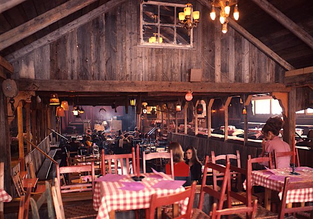 Photo couleur de la salle principale de La Butte. Plusieurs tables avec des nappes à carreaux sont installées devant la scène principale. 
