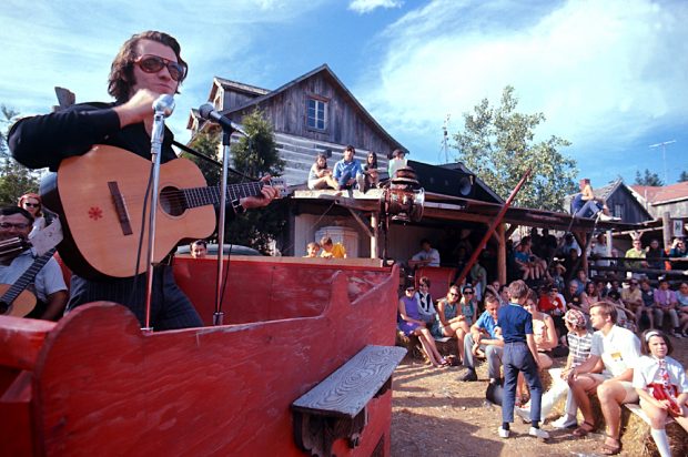 Photo couleur de Jean-Guy Moreau à la guitare, lunettes fumées sur le nez en spectacle à l’extérieur à La Butte devant une foule.