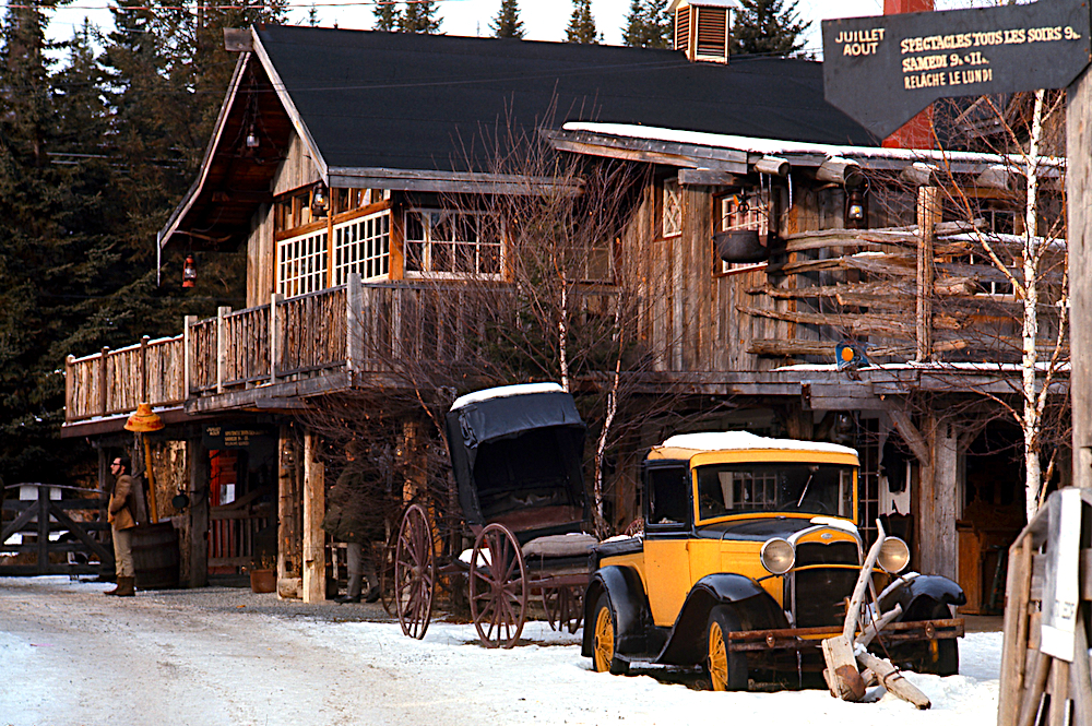 Photo couleur de La Butte en hiver. Les murs de bois et les grandes fenêtres à carreaux sont prédominants. Un ancien camion Ford des années 1930 de couleur jaune est stationné à l’entrée.