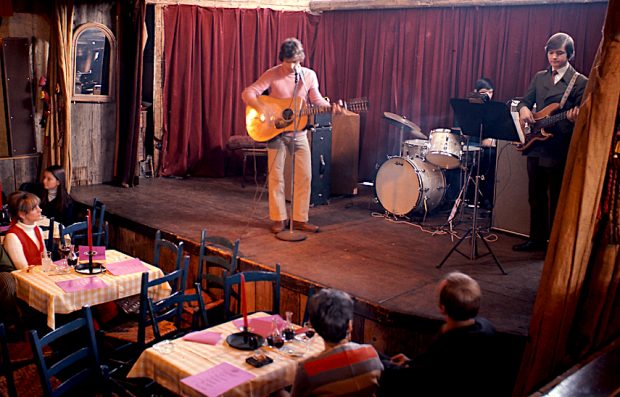 Photo couleur de deux jeunes musiciens sur la scène de La Butte, l’un est debout à la guitare, l’autre est assis derrière une batterie. Des spectateurs attablés dans la salle les écoutent et les regardent.