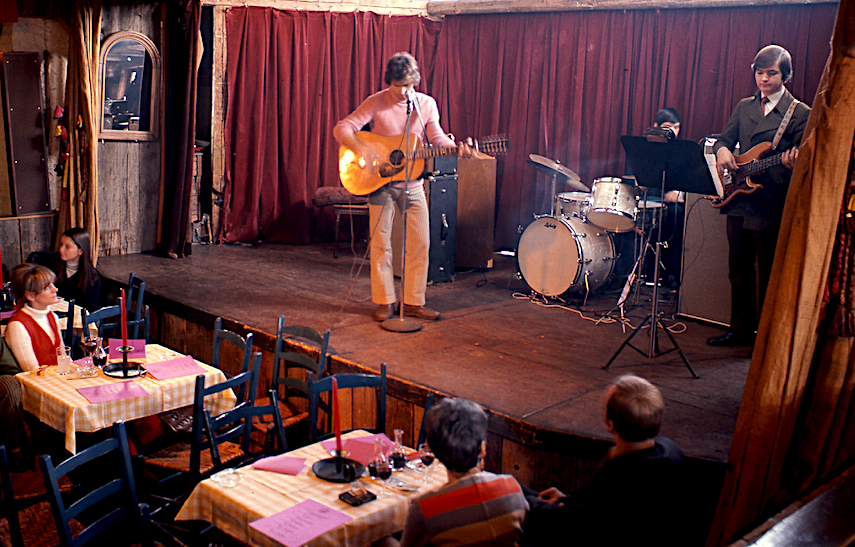 Photo couleur de deux jeunes musiciens sur la scène de La Butte, l’un est debout à la guitare, l’autre est assis derrière une batterie. Des spectateurs attablés dans la salle les écoutent et les regardent.