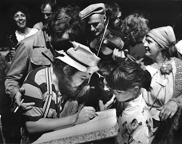 Photo noir et blanc d’amuseurs publics à La Butte. Un groupe d’enfants entoure un clown, un violoneux et Gilles Mathieu.