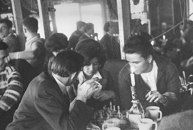 Photo noir et blanc de deux hommes et une femme jouant aux échecs sur une table de la Butte entourés de plusieurs autres personnes.