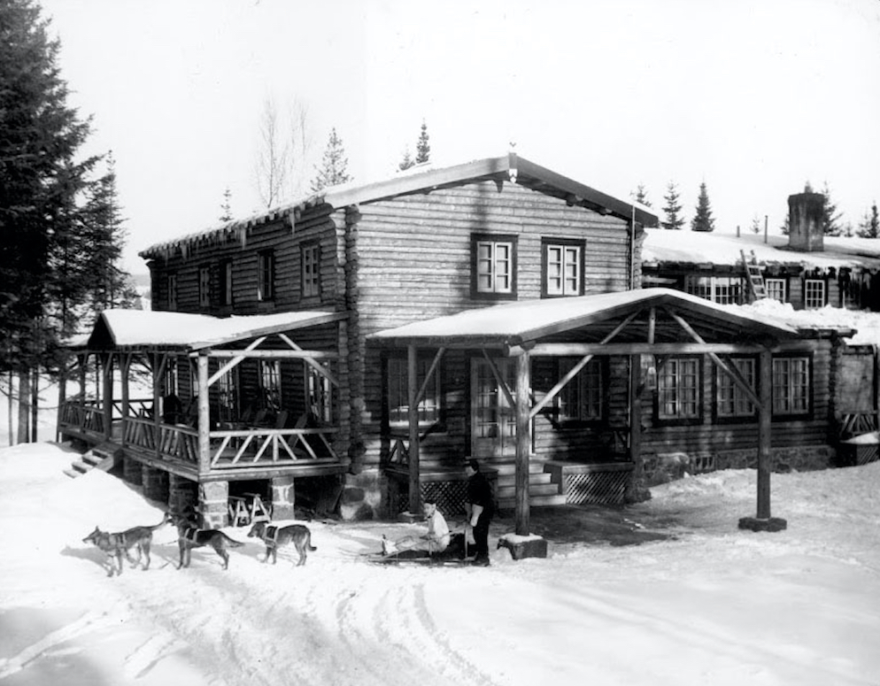 Photo noir et blanc de l’auberge La Sapinière dans la neige à l’hiver 1936-1937. Devant la construction en bois rond, un traineau où sont installées deux personnes est tiré par trois chiens.