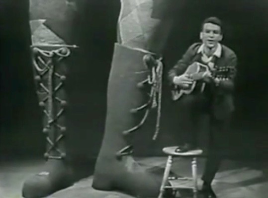 Photo noir et blanc de Claude Gauthier s'accompagnant à la guitare. Une paire de bottes de bûcheron apparaît en avant-plan.
