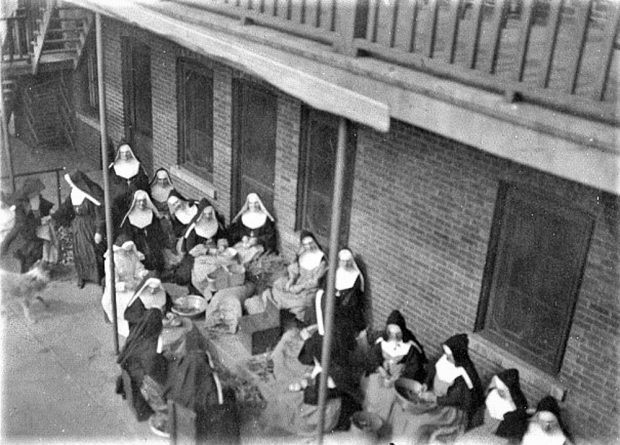 Photo en noir et blanc de plusieurs femmes portant l’habit religieux et le tablier. Elles épluchent des gourganes, adossées au couvent.