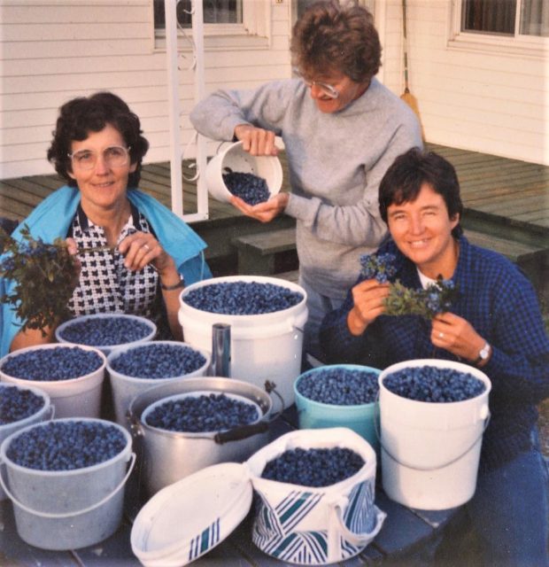 Photo en couleur de trois femmes devant plusieurs contenants de bleuets.