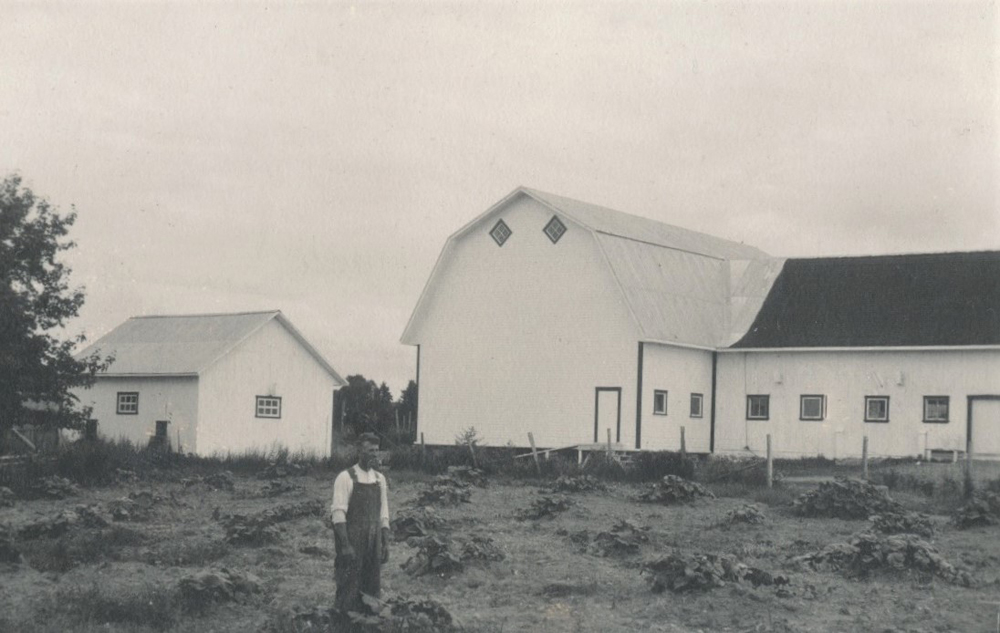 Photo en noir et blanc d’un homme devant la ferme bordée d’un champ.