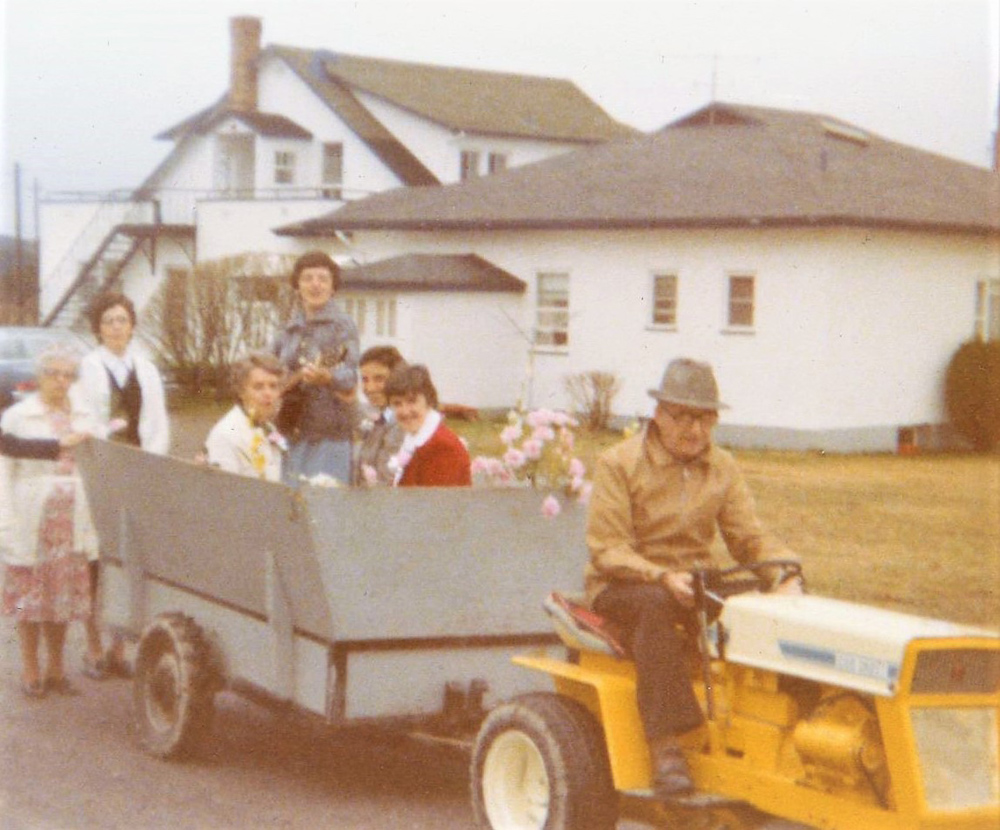 Photo en couleur d’un homme assis sur un petit tracteur jaune traînant une charrette grise où quelques femmes sont assises, dont l’une tient une guitare. Ils se trouvent devant la maison de la ferme.