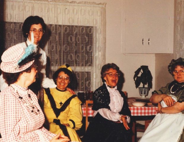 Photo en couleur de cinq femmes dans une cuisine habillées de robes colorées et de chapeaux.