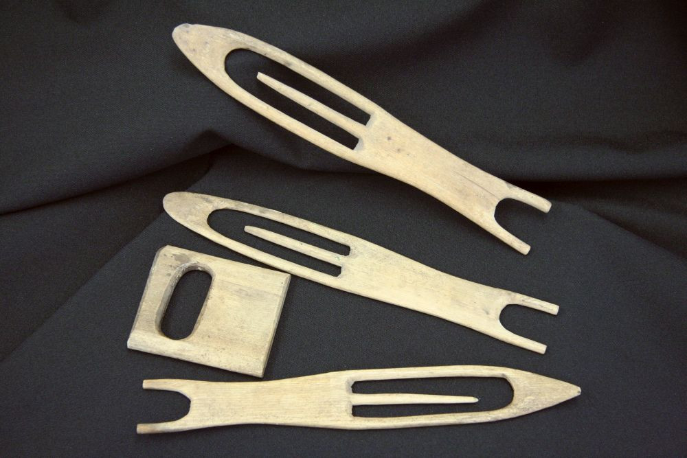 Quatre outils artisanaux en bois mince déposés sur un tissu noir : trois aiguilles en forme d'ogive pour tricoter les filets de pêche et une plaquette carrée servant de gabarit pour la taille des mailles.