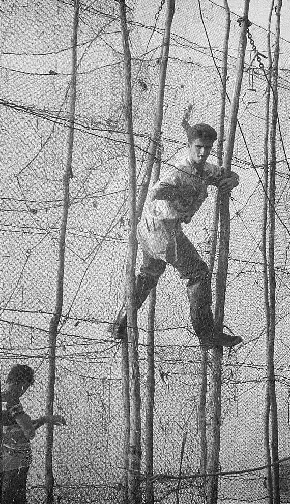 Entre deux rangées de filets de pêche tenus à la verticale par de grandes perches, un homme, s'aidant des perches, se tient en équilibre à environ 3 mètres de haut; photo noir et blanc.