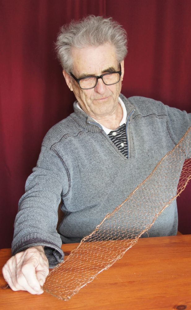 Un octogénaire assis à une table déploie une étroite bande de filet de cuivre entre ses deux mains.