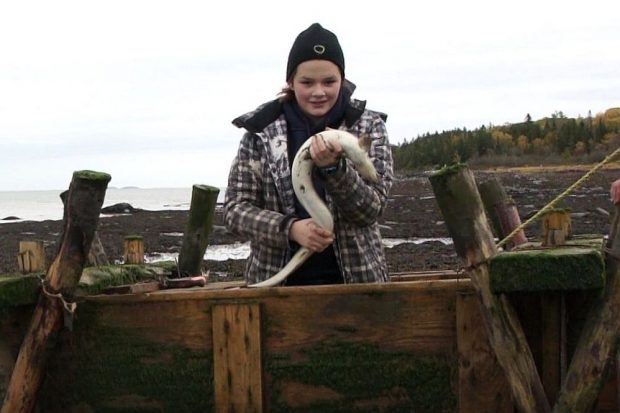 Une adolescente portant une tuque et un manteau est debout dans un coffre en bois servant à garder les anguilles captives dans une pêche. Elle tient une anguille dans ses mains; en arrière-plan, on voit le fleuve et une baie.