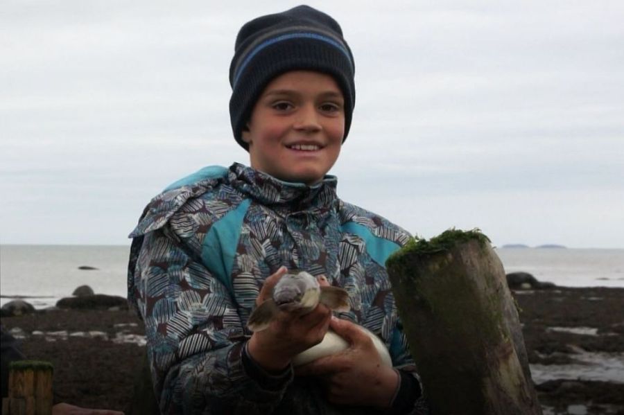 Un jeune garçon, en vêtements d'hiver et vu du torse jusqu’à la tête, tient fièrement une anguille dans ses mains. Il présente la tête du poisson et en arrière-plan on voit le fleuve.