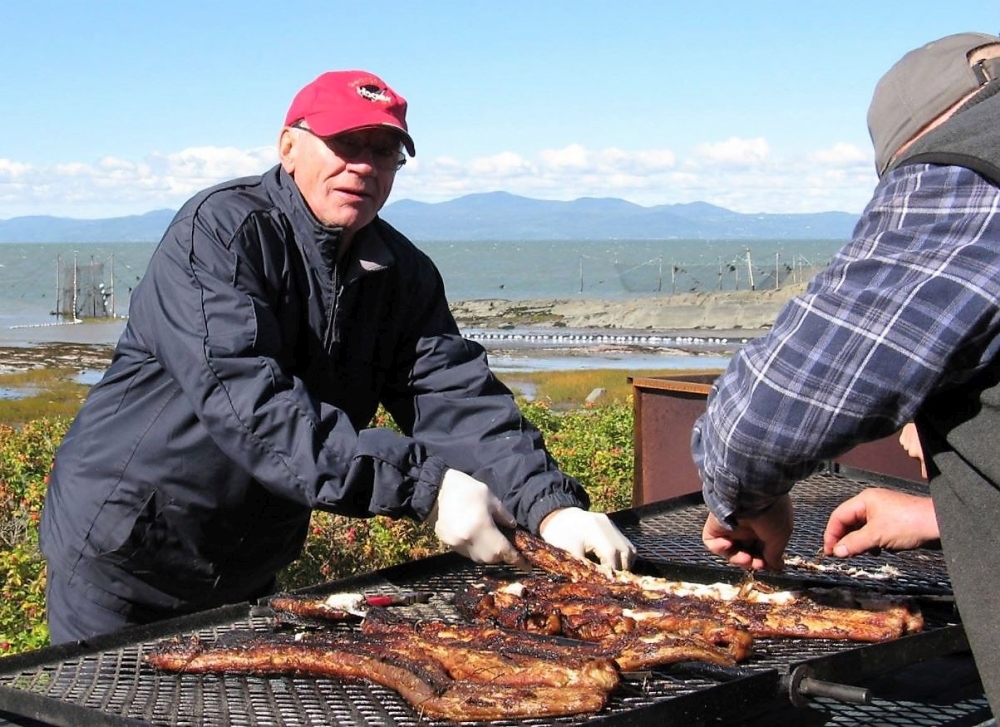 Des anguilles rôtissent sur les grilles d'un B.B.Q. artisanal près du fleuve. Deux hommes s'occupent de la cuisson.