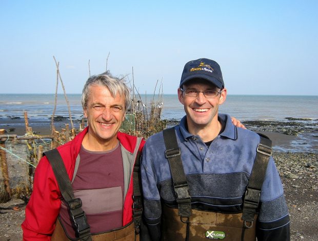 Deux hommes au retour de la pêche à anguilles vus du torse jusqu’à la tête et se tenant amicalement par l'épaule. En arrière-plan, on voit une pêche à anguilles et le fleuve.