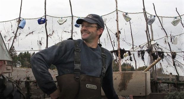 Un pêcheur devant les filets de sa pêche à anguilles vu du torse jusqu’à la tête et ayant sa main droite sur sa hanche.