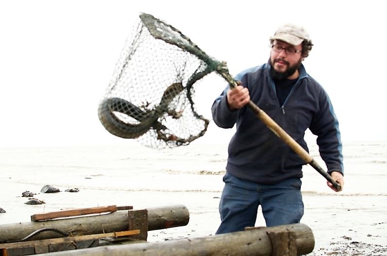 Un pêcheur d'anguilles tient un grand filet attaché à un cadre rigide doté d’un manche et contenant une anguille. On voit le dessus d'un gros coffre en bois au bas de la photo.