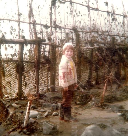 Un petit garçon des années 1970 portant des bottes de caoutchouc, une veste et une tuque de laine pose en souriant devant un filet de pêche à anguilles tenu à la verticale. Il a les pieds dans l'eau.