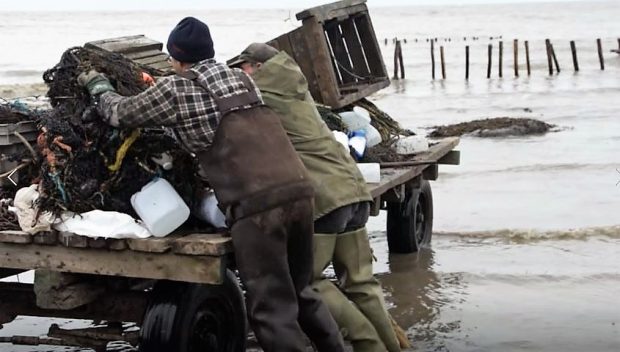 Deux hommes en habit de pêche déposent un filet sur une remorque dont les roues sont dans l'eau montante du fleuve.