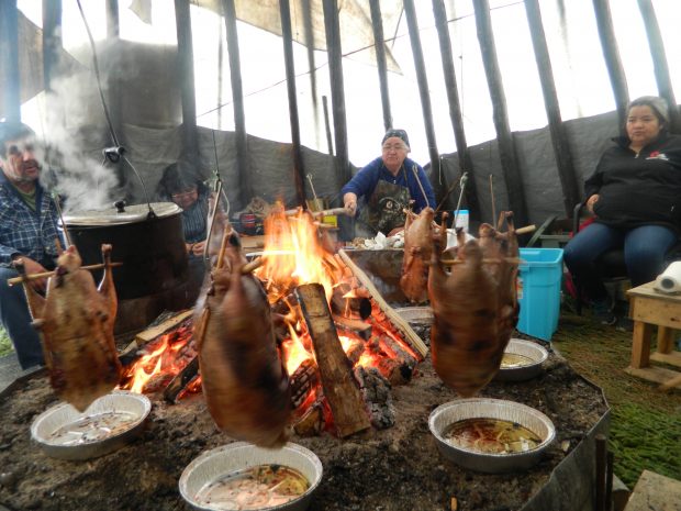 Un homme et deux femmes font braiser une oie sauvage sur le feu dans un tipi