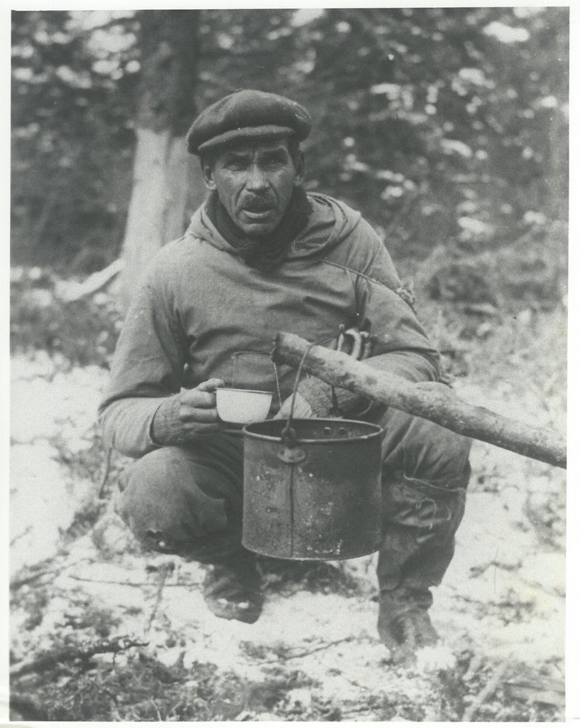 Homme avec un chapeau et une tasse en étain assis près de la forêt. Il a suspendu un seau au-dessus d’un feu pour faire bouillir de l’eau.